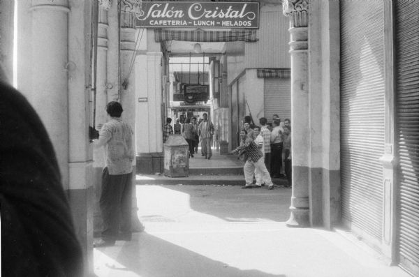 View down a covered Havana sidewalk at the end of the Cuban Revolution.  Small groups of civilians are standing around and walking. A large sign for the Salon Cristal is hanging from the ceiling.