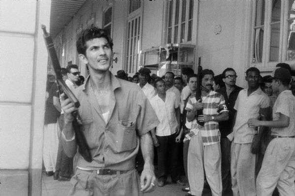 Two of Fidel Castro's 26th of July Movement soldiers wielding guns are standing in front of a crowd next to a building under a covered Havana sidewalk. Taken at the end of the Cuban Revolution in Havana, Cuba.