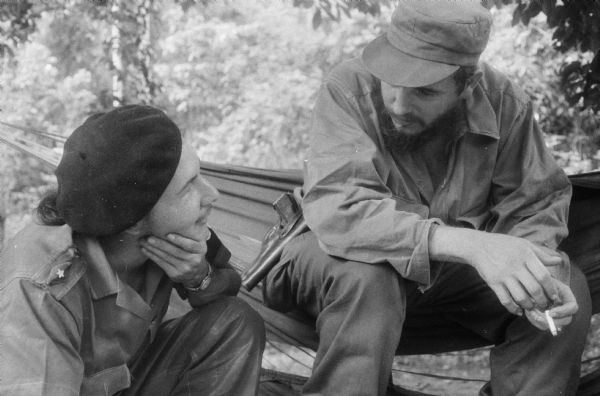 Fidel Castro sits in a hammock smoking a cigarette and speaking with his brother Raul, who is crouching on the ground.