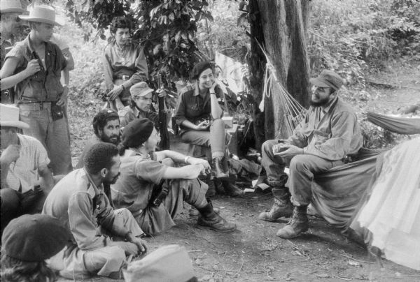 26th of July Movement command headquarters in the Sierra Maestra mountains during the Cuban Revolution.  Fidel Castro sits in a hammock and speaks with Vilma Espin, Raul Castro, Celia Sanchez and several other people.  Raul has just returned from a successful campaign to consolidate revolutionary control of Oriente province.