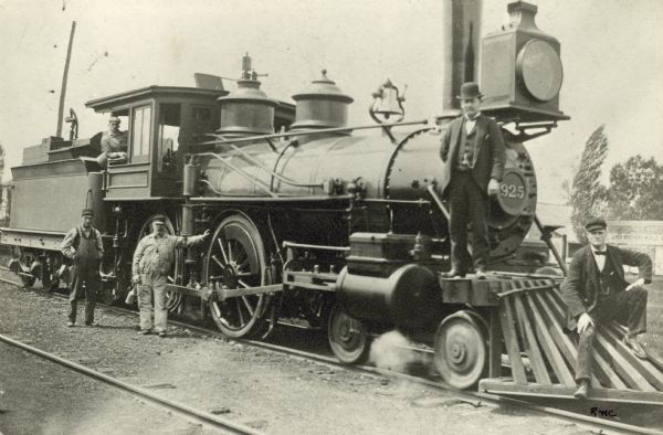Railroad employees pose with locomotive engine no. 925 of the Chicago, Milwaukee & St. Paul Railway. The engine has a long pilot, a large oil headlight, small 6 inch air pump, and two injectors on the right side of the boiler. Fireman James Hart is in the cab window, and engineer Harry Gibson leans on a wheel, holding an oil can. The conductor standing on the pilot beam is probably Hiram Bond, but may be William Schneider.