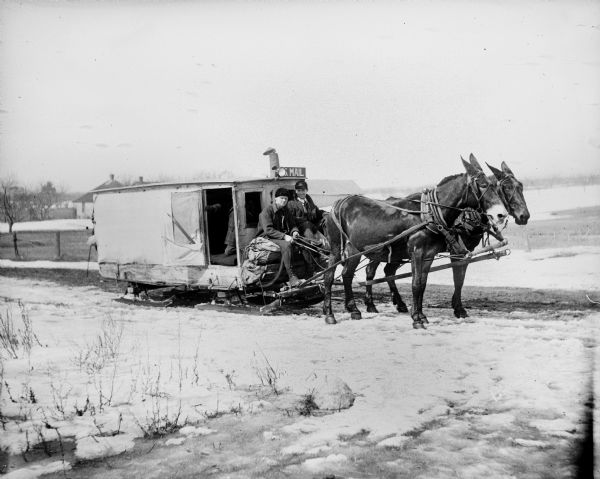 Two men sit at the reins of a horse-drawn United States mail sleigh which ran from Sturgeon Bay to Egg Harbor. Another man is kneeling inside the sleigh near an open door.
