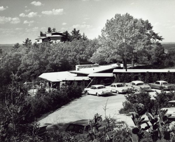 Elevated view over parking lot of House on the Rock. The property is heavily forested with trees, and bushes surround the parking area. There is a long, low building next to the parking area.