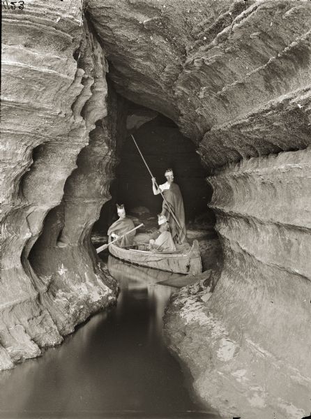 Looking into Boat Cave. Three men dressed as "Indians," one standing on shore holding a spear, two seated in a canoe.