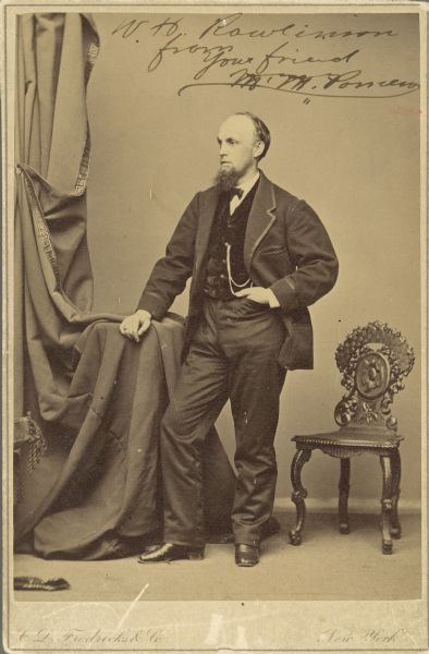 Full-length studio portrait of Marcus Mills Pomeroy standing next to a chair with his hand in his pocket.