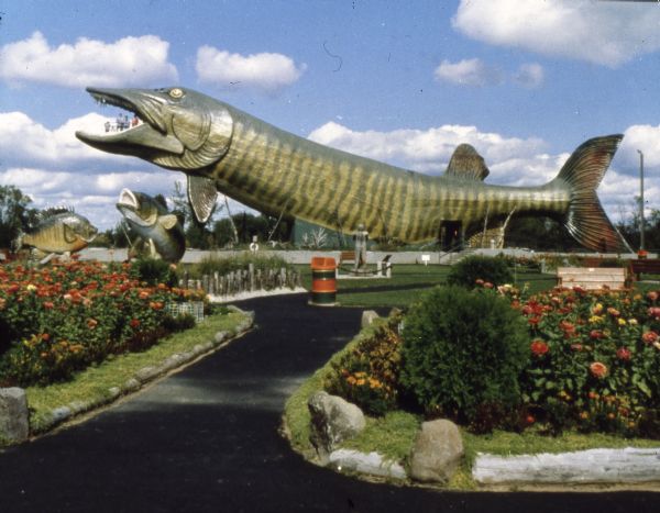 View of the giant walk-through musky and other oversized fish at the National Fishing Hall of Fame. A small group of people is visible in the mouth of the musky. Landscaping with shrubs and flowers appears in the foreground.