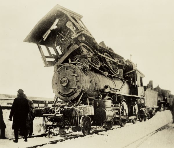 View of the aftermath of a train wreck that left one locomotive on top of another. A small group of men stand in the snow along the tracks near the wreck. The bottom locomotive is #823.