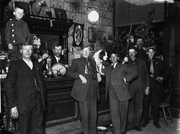 Group of men gathered at the bar in McGlaughlin and Reynolds Bros. Saloon. There is a young boy standing on the bar, as well as a dog sitting on the bar. One man is holding a gun. A mounted deer head is mounted above the bar. A bartender with a moustache stands behind the bar holding a decorative serving tray with the name of the bar in one hand, and in the other a shot glass on top of a cut glass decanter.