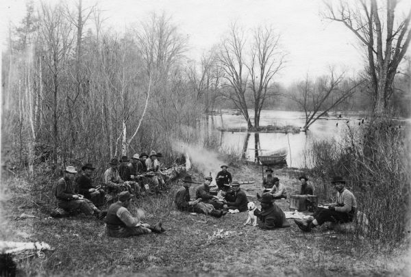 Log driving crew sitting down for lunch on the bank of a river (probably the Trap River or Rib River). There is a batteau at the water's edge, and one man holds a dog.