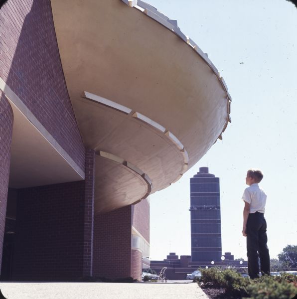 Boy standing below the Golden Rondelle with the Johnson Wax Research Tower in the background. Workers on scaffolding can be seen on the tower in the distance.