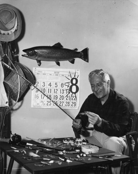 Governor Warren Knowles holding a fishing pole and a fly. He is seated at a table covered in fishing tackle and additional fishing gear hangs on a rack at left. There is a stuffed trout on the wall above a May 1965 calendar page with an oversized 8 to show the opening date of fishing season in Wisconsin.