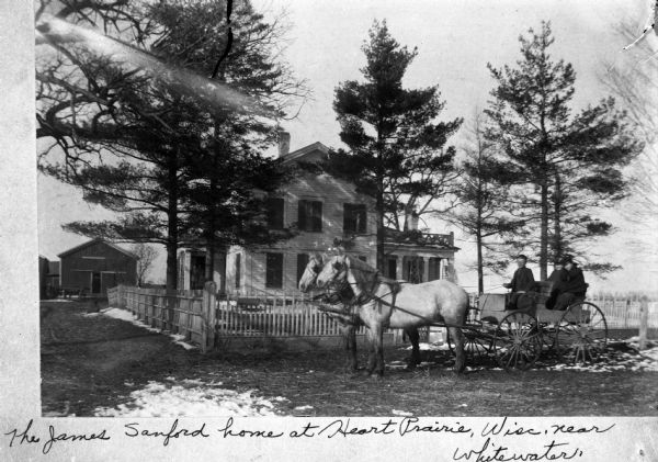 Young man and two women in a horse-drawn wagon in front of the fenced yard of the James Sanford home. There is a small patch of snow on the ground in front of the horses.