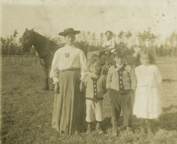 Left to right, Maria, Fred, Oscar, and Alma Getto standing in a field. Henry Getto is in a horse-drawn wagon in the background.