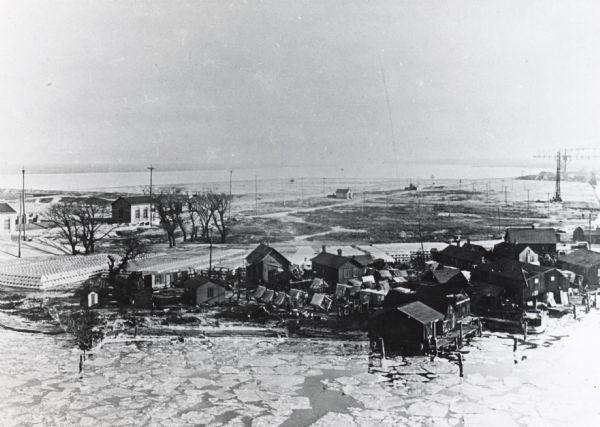 Elevated view of Jones Island fishing community in Milwaukee Harbor. Several shacks are visible as well as many fishing nets on net rollers. Ice on the water suggests it was winter or early spring.