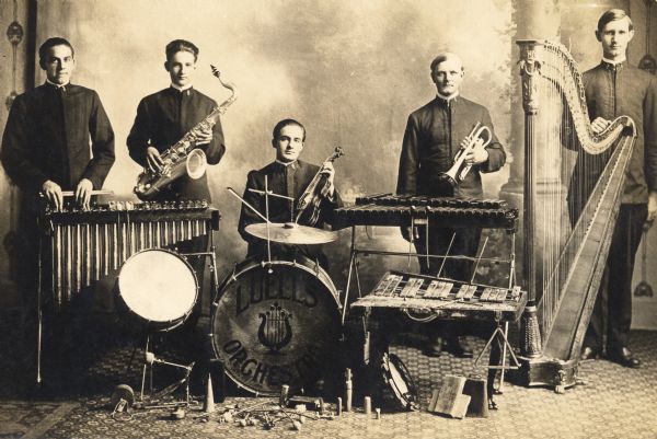 Indoor group portrait in front of a painted backdrop of Luell's Orchestra consisting of five men. Each member holds an instrument and there are several other musical instruments positioned around the scene. Instruments included are: vibraphone, saxophone, drum kit, violin, xylophone, trumpet, harp, cowbell, wood blocks and tambourine.