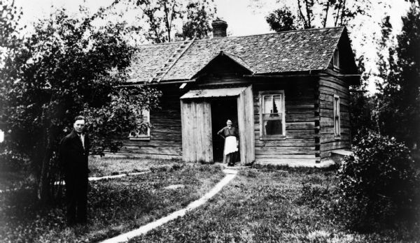 Outdoor portrait of Henry Getto standing in the yard, and Maria Getto standing in the doorway of their log home. There is a ladder on the roof.