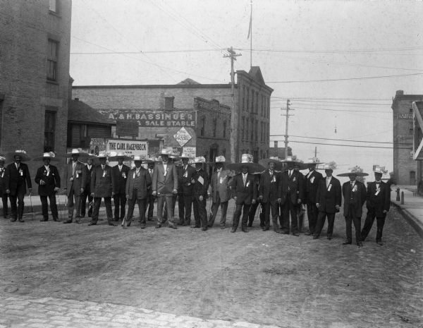 Outdoor group portrait of the Racine Chapter of the Benevolent and Protective Order of Elks wearing suits, hats with oversized brims with large flower decorations, and buttons and ribbons on their lapels. Each man holds a cane and they are standing together in the middle of a street.