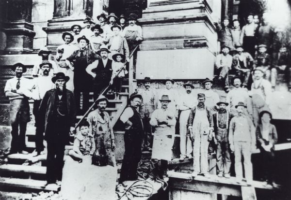 Construction workers are posed in front of the partially-built St. Josaphat's Church on scaffolding and wooden steps near large columns. There are two children standing with the men on the left side.