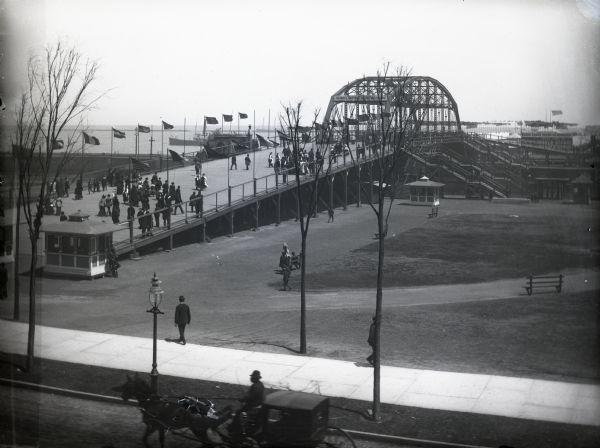 Elevated view of the Illinois Central World's Fair Transportation Center at Van Buren Street. Several pedestrians cross a bridge leading to trains and lake steamers. A horse-drawn carriage passes along a street in the foreground.