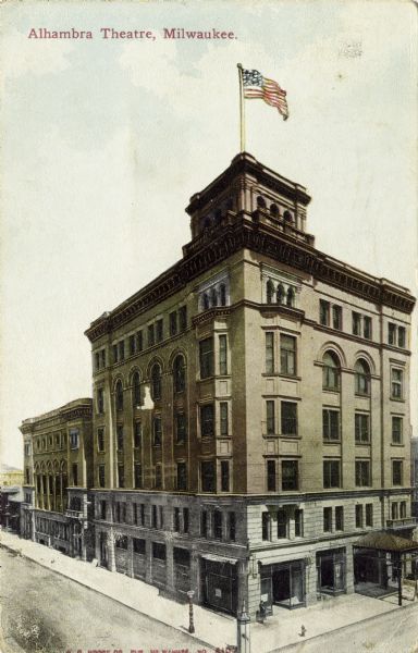 Elevated view of the Alhambra Theatre Building on the corner of W. Wisconsin Avenue and N 4th Street. A U.S. flag flies from the top of the building. Caption reads: "Alhambra Theater, Milwaukee."
