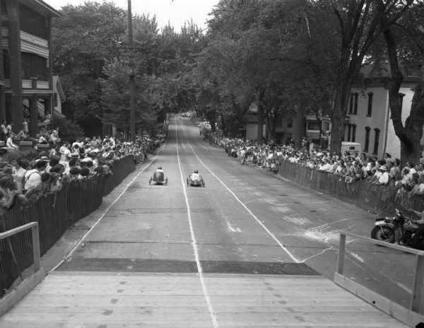 Two racers streak down the soap box derby track on E. Gorham Street as a large crowd watches from behind a fence along the sidewalk.
