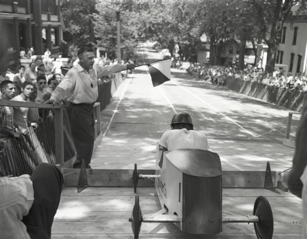 View from behind of a soap box racer in the starting gate at the top of the E. Gorham Street hill between N. Butler and N. Pinckney Streets. A man with a flag waits to start the race as a large crowd watches from behind a fence along the sidewalk.