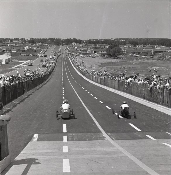 Soap box racers head down a hill on S. Midvale Boulevard as a large group of spectators are watching from behind a fence. 1953 was the first year Madison's soap box derby was run on S. Midvale Boulevard.