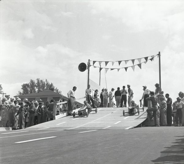 Two soap box racers leave the starting gate on their way down the hill on S. Midvale Boulevard. A few spectators can be seen on both sides of the starting ramp. 1953 was the first year Madison's soap box derby was run on S. Midvale Boulevard.