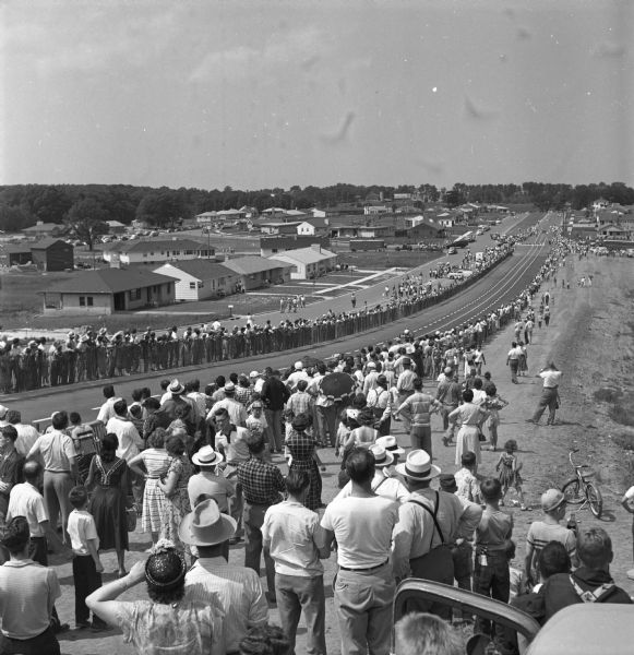 View down the hill at the Madison Soap Box Derby on South Midvale Boulevard during a soap box derby race. One car is visible on the track and a large crowd is watching from behind a fence. 1953 was the first year Madison's soap box derby was run on S. Midvale Blvd.