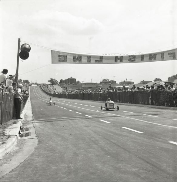 Two Madison soap box derby contestants are racing toward the finish line on South Midvale Boulevard as a large crowd watches from behind a fence. The 1953 race was the first held on South Midvale Boulevard in Madison.