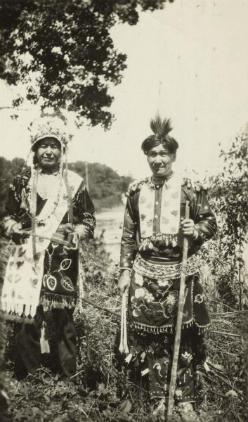 Outdoor portrait of Peter Wolfe, chief of Lac Courte d'Oreilles band of Ojibwa (Chippewa) Indians, with his son William Wolfe, who was Secretary of Lac Courte d'Orielles. Both men wear traditional, beaded garments. Peter holds a pipe and William holds a decorated staff.