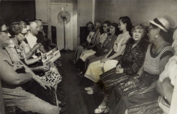 Men and women seated on benches along both walls of Kenneth Crook's Uranium Tunnel in Lone Rock. Note the uranium pads visitors hold in their laps or on their feet. Padding on the floor also contains uranium ore. One woman reads and another knits while absorbing the low grade radiation. There is a floor fan at one end of the tunnel.