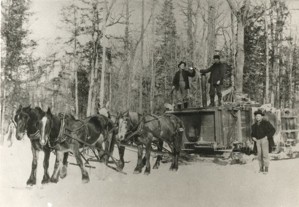 Two men standing on a water tank sleigh used for icing logging roads. The sleigh is pulled by four horses. Foreman Mike Baltus stands in the snow at right. One of the men standing on the water tank is holding a bottle. A large saw is also on top of the tank.