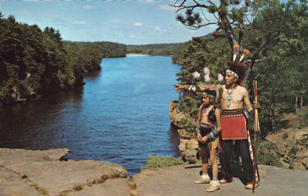 An Indian man and boy in native garb stand on High Rock with the Wisconsin River in the background. The man is pointing towards the left and holding a bow and arrow.