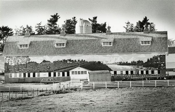 Pastoral panoramic painting on the side of a barn. Two people on scaffolding can be seen working on the mural at right. A silo juts above the barn. There is a station wagon parked in front near a fenced area on the left, and on the right is a tractor hitched to a wagon a group of Holstein cows.
