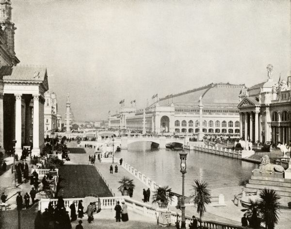 View of the canal area of the Columbian Exposition grounds looking north from near the front of the Machinery building (at left). The South canal is in the foreground with bridges crossing it. The Agriculture Building is seen at right and the Manufacturers and Liberal Arts Building is in the center. A drinking water station can be seen at the bottom center of the image. Many people are walking around the grounds and observing the scenery. A number of statues can be seen stationed around the water and on buildings.