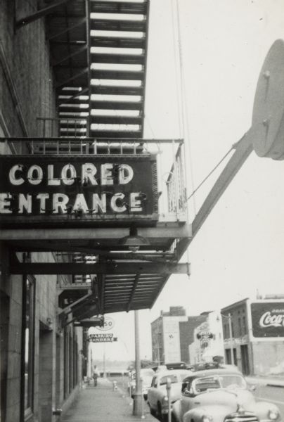 Exterior view of the segregated entrance for African-Americans at Malco Theater. There is a fire escape on the side of the building. Several cars are parked at meters along the curb. There is a Coca-Cola advertisement on a building across the street.