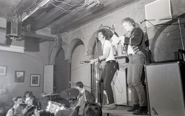 Rock trio, "The Ox," performing on stage. The guitarist and bassist are on an elevated platform next to a stack of amplifiers, while the drummer is set up on the floor. A few audience members are sitting at tables.