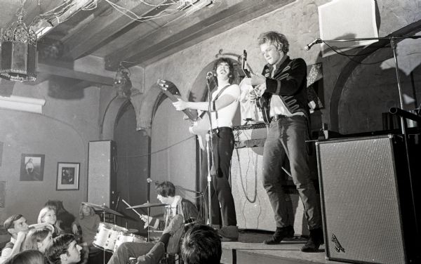Rock trio, "The Ox," performing on stage. The guitarist and bassist are on an elevated platform next to a stack of amplifiers while the drummer is set up on the floor. A few audience members are sitting at tables.