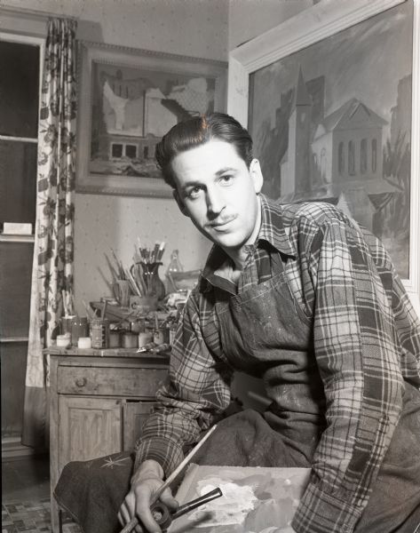 Portrait of artist, Lester Bentley, wearing an apron and sitting in his studio holding a brush, a pipe and a painters palette.