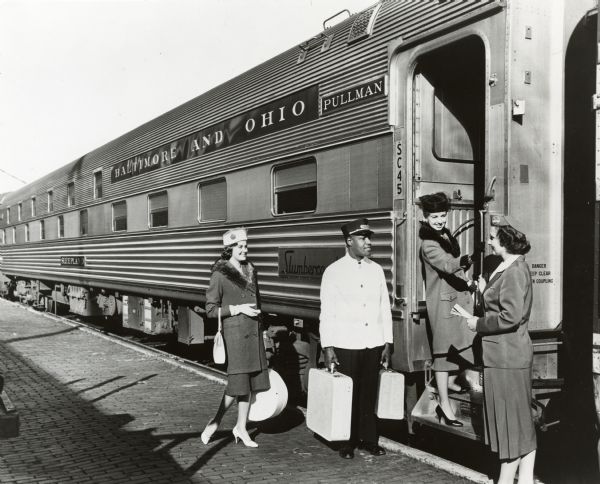 Two women boarding the Slumbercoach car on a B&O Railroad passenger train. A Pullman porter carries two suitcases as a stewardess greets customers. .