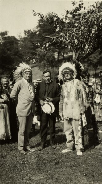 Outdoor portrait of former boxer, Tommy Gibbons, Father Gordon, and Governor Fred R. Zimmerman at a Ho Chunk (Winnebago) ceremony. Zimmerman is wearing a buckskin, and both he and Gibbons are wearing headdresses. The photograph was probably taken at a ceremony in Frost's Woods, Blooming Grove, Wisconsin.