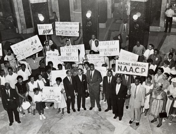Large group of protesters in the Wisconsin State Capitol rotunda with many carrying signs in favor of Civil Rights for African-Americans. One man holds a sign for the Racine Branch of the National Association for the Advancement of Colored People (NAACP). Isaac Coggs of Milwaukee stands in front at center and Lloyd Barbee is to the left of him beneath a sign reading "Democracy Not Hypocrisy."