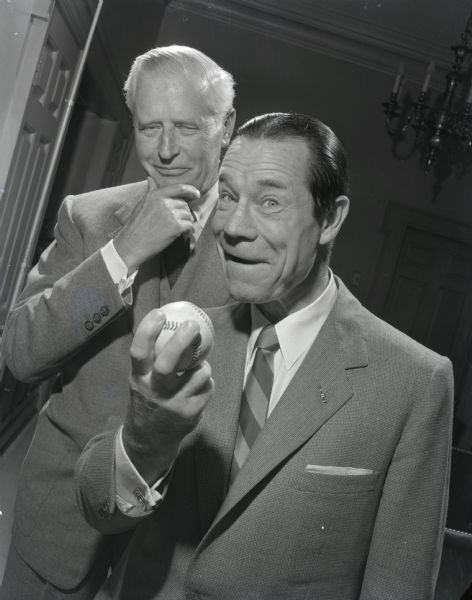 Comedic actor Joe E. Brown holding a baseball, with an unidentified man standing behind him..