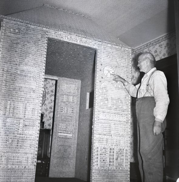 Man pointing to a wall clock on the exterior of a model building completely papered with postage stamps.