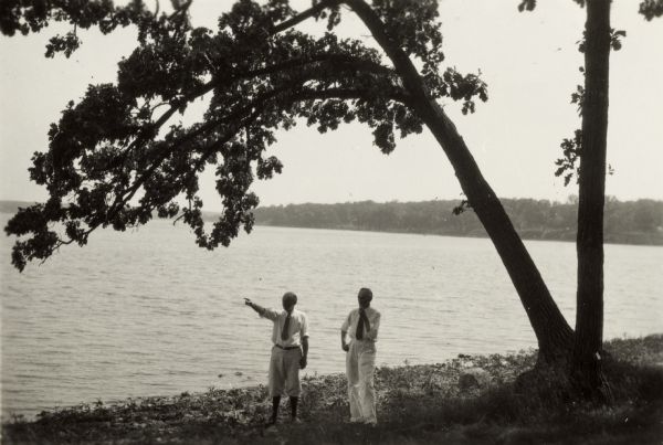 Unidentified man pointing, and Charles E. Brown standing to his side, are beneath a tree on the shoreline of a lake (possibly Lake Monona).