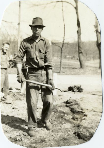 Man in work clothes holding a shovel loaded with dirt while another man watches in the background. The men were Civilian Conservation Corps workers at Devil's Lake State Park Camp.