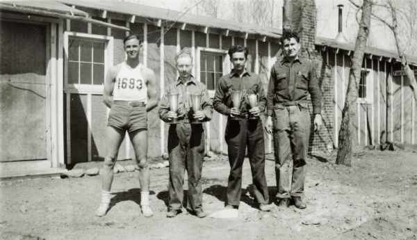 Russ Edlerkin, Army Chief Clerk in shorts and tank top; George Arthur Weidner, Army Chief Clerk holding two trophy cups; unknown F.S. Clerk holding two trophy cups; and unknown F.S. Assistant Clerk standing outside the barracks at the Jump River Civilian Conservation Corps camp (no. 1693 later changed to 3651).