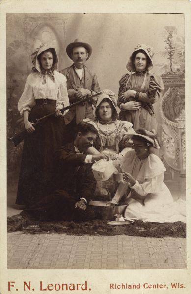 Informal studio portrait of Ada James holding a gun, and her father, David G. James in a hat, with four other people dressed in pioneer-type costumes. A woman at right stands holding a cat, and a man pours from a ewer into a tin cup held by a woman seated at right.