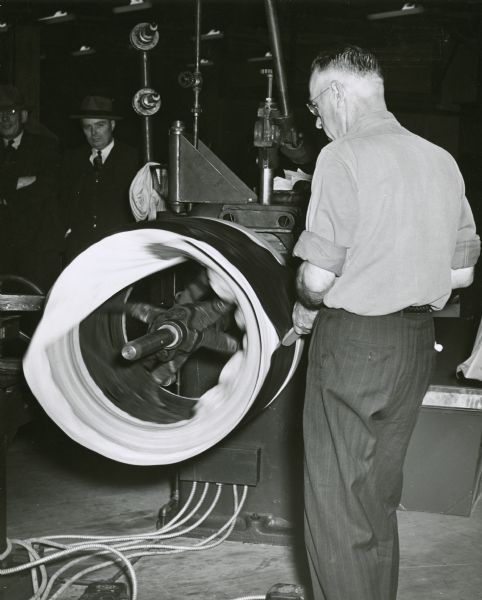 Veteran tire builder Bill McNair works on a pocket while constructing a heavy service truck tire. Two men wearing suits and hats stand in the left background.
